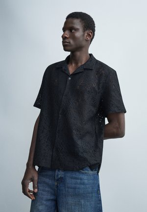 Άνδρας με σκούρο δέρμα που φοράει μαύρο πουκάμισο με κοντά μανίκια και σχέδιο με κουμπιά και μπλε τζιν, στέκεται μπροστά σε απλό ανοιχτό γκρι φόντο.