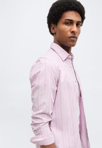 Νέος άνδρας με άφρο κούρεμα που φοράει ένα ανοιχτό ροζ πουκάμισο με μακριά μανίκια και λεπτές κάθετες ρίγες, στέκεται μπροστά σε ένα απλό φωτεινό φόντο.