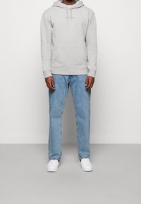 Sudadera gris claro con capucha, bolsillo frontal y logo discreto. Combinada con vaqueros de corte holgado en azul claro y zapatillas blancas.