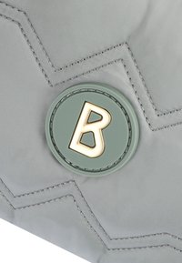 Grå tygväska med en chevron-design, med en grön cirkulär logotyp och ett guld "B" i mitten. Slät textur.