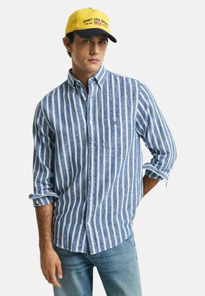 Camicia a righe blu e bianche con taschino sul petto, maniche lunghe arrotolate fino ai gomiti; abbinata a un cappellino da baseball giallo e blu navy.