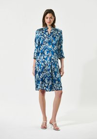 Abito in satin blu con stampa floreale, lunghezza al ginocchio, con collo a camicia e maniche a tre quarti. Indossato con sandali bianchi con lacci.