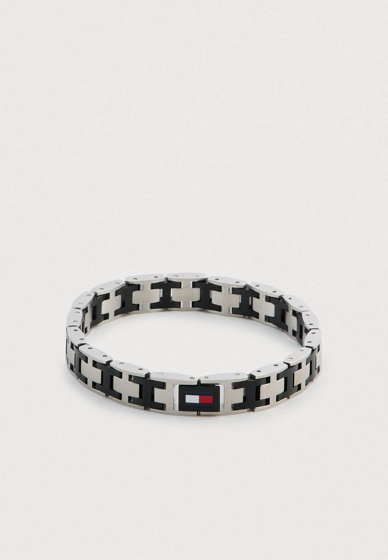 Metalowa bransoletka z naprzemiennymi srebrnymi i czarnymi ogniwami, z prostokątnym zapięciem ozdobionym logo w kolorach czerwonym, białym i niebieskim.