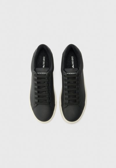 Black sneakers made of smooth material with six eyelets, flat black laces, and a contrasting white rubber sole. Logo visible on insole.