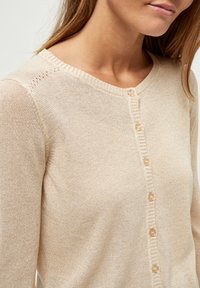 Gaišbeige cardigan, kas izgatavots no mīksta, teksturēta trikotāžas auduma, ar apaļu kakla izmēru, dekoratīvu pogu aizdari un smalku detalizāciju tuvumā pleciem.