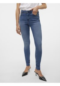 Femme portant un jean skinny bleu denim ajusté et des escarpins noirs à bout pointu, avec une main dans la poche, sur fond blanc.