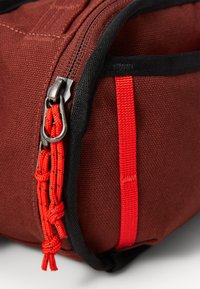 Sac en tissu marron avec une fermeture éclair noire, équipé d'un tirant en paracorde rouge et d'une sangle d'accent rouge. Matériau texturé avec coutures robustes.