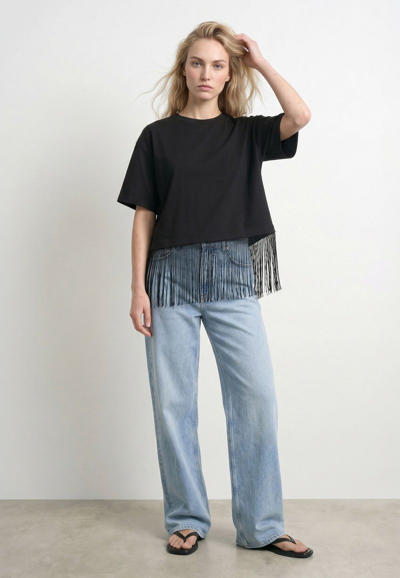 Femme blonde portant un haut noir à manches courtes avec des franges, un jean large bleu clair et des tongs noires, debout devant un fond uni.