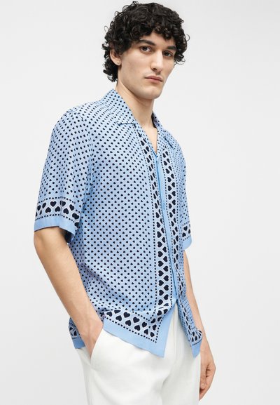 Νέος άνδρας με σγουρά μαλλιά, φοράει ανοιχτό μπλε κοντομάνικο πουκάμισο με σχέδια σκούρου μπλε καρδιές και λευκό παντελόνι, τα χέρια στις τσέπες.