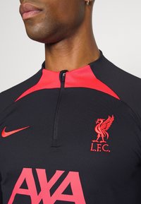 Musta urheilupaita punaisilla yksityiskohdilla, jossa on vetoketjullinen kaulus, Nike-logo, Liverpool FC:n vaakuna ja rohkeat vaaleanpunaiset "AXA" -tekstit.