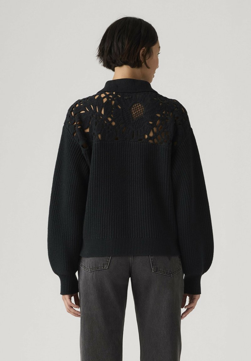 Vrouw die een zwarte gebreide trui draagt met een open kanten patroon op de bovenrug, gecombineerd met donkergrijze spijkerbroek, met de rug naar de camera toe.