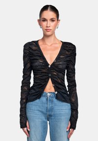 Schwarze, durchsichtige Langarmbluse mit strukturiertem Muster, V-Ausschnitt und Knopfverschluss. Kombiniert mit hochgeschnittenen blauen Jeans.
