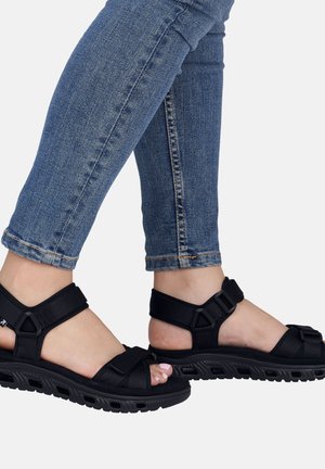 Pieds portant des sandales de sport noires avec des brides réglables et un vernis à ongles rose visible, associés à un jean skinny bleu retroussé.