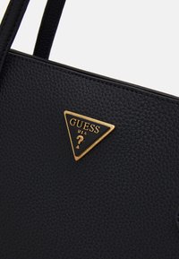 Czarna torebka o fakturowanej powierzchni z wyraźnym złotym trójkątnym logo z napisem „GUESS USA”. Torba ma solidne paski i elegancki design.