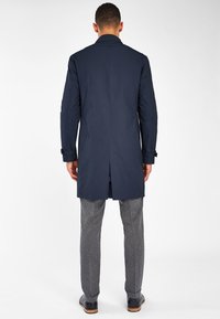 Abrigo trench de color azul marino, hecho de una tela suave, con un corte recto, apertura en la parte posterior y puños abotonados, combinado con pantalones grises texturizados.
