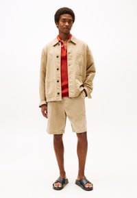 Beige cotton jacket with black buttons and pocket, red mesh shirt, beige shorts, and black slide sandals. Neutral colors, casual design.