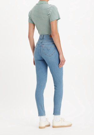 Jeans en denim bleu taille haute avec une coupe slim, dotés d'un design de poche arrière en forme de cœur. Associés à un polo rayé vert et blanc.