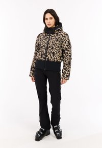 Luipaardprint pufferjack met een zwarte capuchon, aansluitende zoom en ritsluiting, gecombineerd met zwarte skibroeken en zwarte skischoenen.