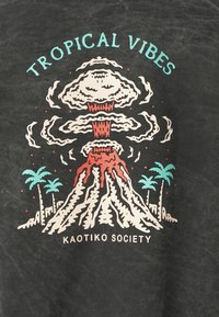 Czarny t-shirt z kolorową grafiką wybuchającego wulkanu z palmami, opisaną jako „Tropical Vibes” i „Kaotiko Society”.