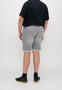 Hombre vistiendo pantalones cortos de mezclilla gris claro remangados, camiseta negra de manga corta, calcetas negras y zapatillas deportivas negras con suelas de goma, de pie contra un fondo plano.