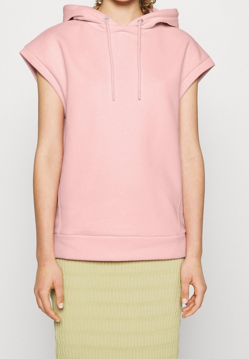 Sweat à capuche sans manches rose en tissu doux avec une poche avant et des cordons de serrage ajustables ; accompagné d'une jupe texturée vert clair.