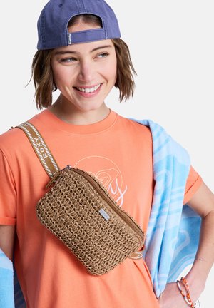 Young woman wearing a blue cap backward and orange T-shirt, carrying a woven crossbody bag, with a blue towel draped over her shoulders.