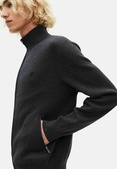 Ciemnoszara rozpinana bluza z wysokim kołnierzem, bocznymi kieszeniami i ściągaczami, z subtelnym logo na klatce piersiowej. Gładka tekstura.