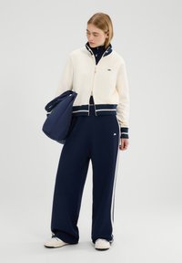 Chándal azul marino con acentos crema, que incluye una chaqueta con cremallera y puños acanalados y cuello alto, combinada con pantalones de pierna ancha y un bolso a juego.