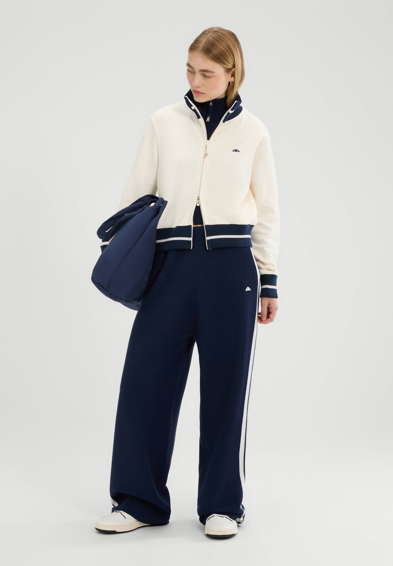 Chándal azul marino con acentos crema, que incluye una chaqueta con cremallera y puños acanalados y cuello alto, combinada con pantalones de pierna ancha y un bolso a juego.