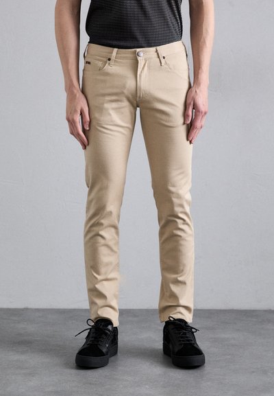 Beżowe spodnie slim-fit charakteryzują się gładką fakturą, pięcioma kieszeniami, metalowym zapięciem na guzik i są zestawione z czarnymi trampkami.