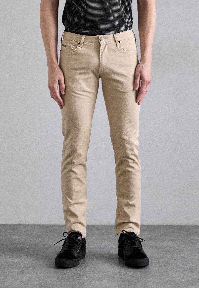 Bež hlače slim fit imaju glatku teksturu, dizajn s pet džepova, metalno kopčanje i kombinirane su s crnim tenisicama.
