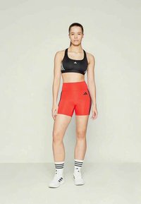 Žena stojí oblečená v čiernej športovej podprsenke, červených vysokých šortkách, bielych ponožkách s čiernymi pruhmi a bielych teniskách na jednoliatom pozadí.