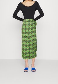 Jupe enroulée verte à motifs avec des motifs noirs en forme de coquillage, associée à un haut noir à manches longues et des sandales ouvertes bleues.