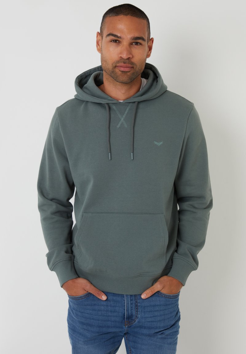 Groene hoodie met een voorzak, trekkoord capuchon en subtiele logo-accent. Zachte stof met een gladde textuur, gecombineerd met blauwe jeans.