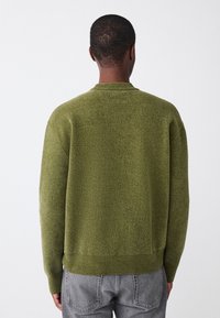 Olivzaļš, teksturēts adīts džemperis ar apaļu kakla izgriezumu, ribots aproču un apakšmalas apdare. Valkāts kopā ar gaisīgi pelēkiem, brīvā piegriezuma džinsiem.