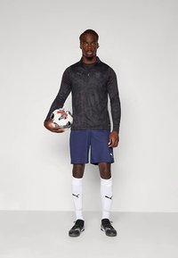 Vyrų futbolininkas, dėvintis juodą ilgomis rankovėmis marškinėlį ir tamsiai mėlynas šortus, laikantis futbolo kamuolį viena ranka, stovintis prieš vientisą foną.