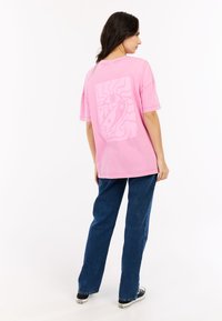 Roze t-shirt met korte mouwen met een grote witte print op de achterkant, gecombineerd met blauwe denim jeans en zwarte sneakers. Casual pasvorm en relaxte stijl.