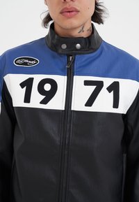 Läderjacka med blå, svarta och vita sektioner. Stora svarta siffror "1971" på framsidan, med dragkedja och tryckknappskrage.