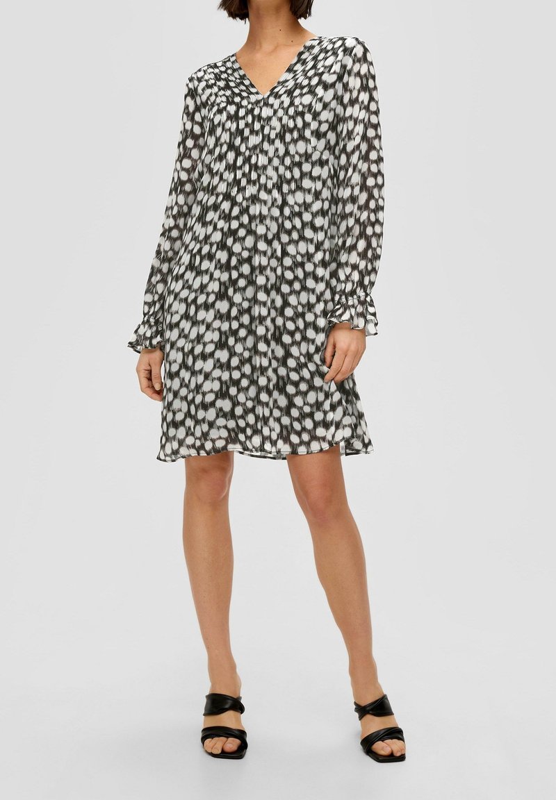 Femme portant une robe noire et blanche à manches longues, longueur genou, avec un col en V et un motif abstrait à pois, associée à des sandales noires ouvertes à l'avant.