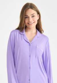 Lila långärmad pyjamatopp med knäppning, kavajkrage och bröstficka med kontrasterande piping längs kragen.