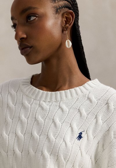 Λευκό πουλόβερ με πλέξη καλώδιο, στρογγυλή λαιμόκοψη και ριμπ μανσέτες, με μικρό ναυτικό λογότυπο στο στήθος. Χρυσά σκουλαρίκια με λεπτομέρειες που μοιάζουν με κοχύλια.