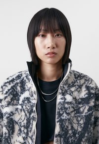 Fleece jacket in black, white, and gray with a textured, fluffy surface. Features a high collar, zipper, and a triangle logo on the chest.