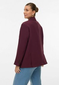 Bordeaux blazer i en skræddersyet pasform med notchede slag, skulderpuder og knappede manchetter. Kombineret med lyseblå jeans.