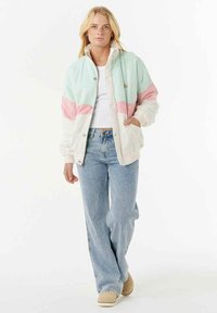 Donna che indossa un giubbotto colorato in pastello, una maglietta corta bianca, jeans azzurri larghi e scarpe beige, che cammina verso la macchina fotografica su uno sfondo bianco.