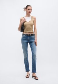 Brązowy prążkowany top na ramiączkach nałożony na białą koszulę, zestawiony z jasnoniebieskimi jeansami o prostych nogawkach, czarnym paskiem z okrągłymi ozdobami i czarnymi sandałami.