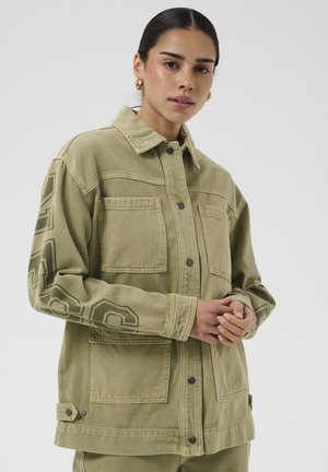 Femme aux cheveux foncés et boucles d'oreilles créoles dorées portant une veste oversize vert olive avec de grandes poches et un texte sur la manche, mains jointes.