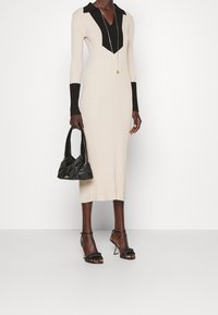 Abito a maglia beige a coste con colletto e polsini neri, abbinato a una borsa nera trapuntata e tacchi neri con cinturini. Collana con pendente in argento inclusa.