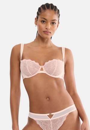 Kobieta z zaplecionymi włosami, ubrana w jasnoróżowy koronkowy biustonosz i pasujące przezroczyste majtki, stoi na tle gładkiego białego tła.