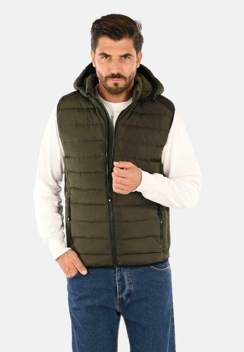 Gilet imbottito verde scuro con chiusura a zip, cappuccio e tasche laterali con zip, indossato sopra una maglietta bianca a maniche lunghe e jeans blu.