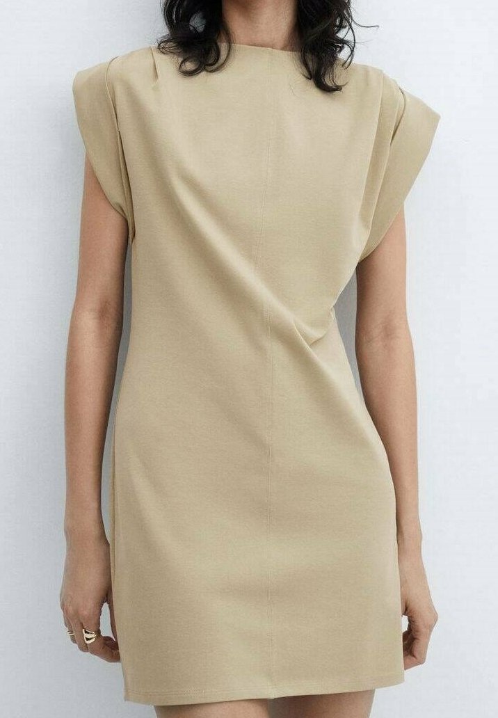 Femme portant une mini-robe beige sans manches avec des épaules structurées, debout devant un fond blanc uni.